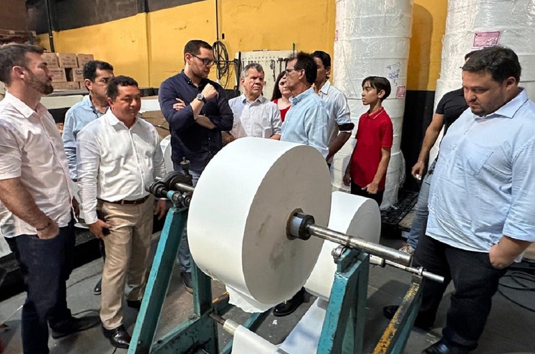 Imagem mostra pessoas em pe vendo uma linha de produção de papel para guardanapos