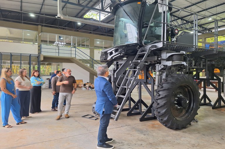 A imagem mostra um grupo de pessoas em um galpão industrial observando uma grande máquina agrícola elevada sobre suportes. Um homem de terno azul está na frente, enquanto outro, de camiseta marrom, parece explicar algo. O ambiente tem estrutura metálica, iluminação natural e um andar superior com paredes de vidro.