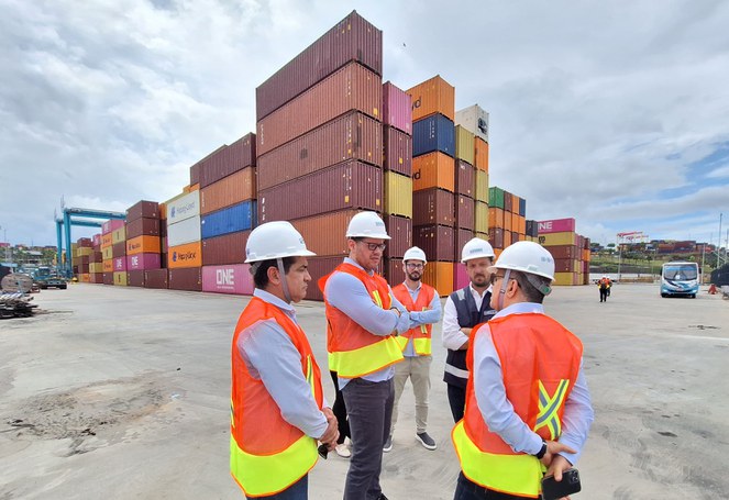 Foto mostra grupo de pessoas com capacete branco e contêineres ao fundo, durante visita a um terminal de cargas