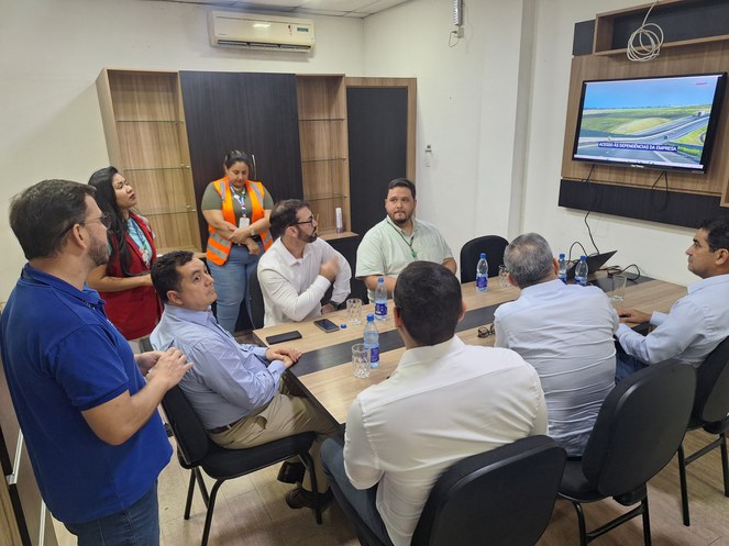 Foto mostra homem de blusa azul fazendo uma explicação a um grupo de pessoas em uma sala fechada e com um telão de apoio