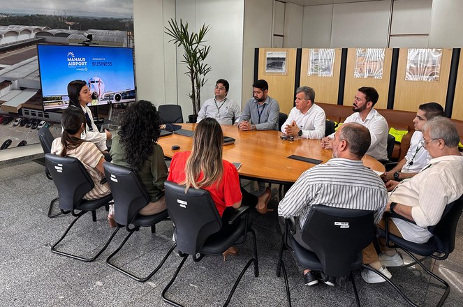 Foto mostra grupo de pessoas sentadas à mesa numa reunião, com um telão ao fundo