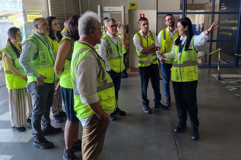 Foto mostra mulher levantando o braço e apontando para uma direção, enquanto faz uma explicação ao um grupo de pessoas vestidas jaquetas verdes