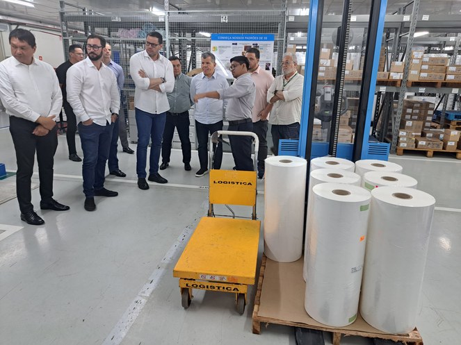 Foto mostra homem de blusa azul e óculos gesticulando, durante explanação para um grupo de pessoas