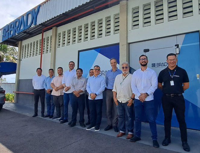 Foto mostra um grupo de pessoas posando para foto em frente a uma fábrica