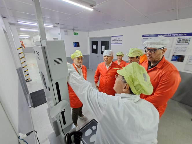 Foto mostra mulher vestida de jaleco e boné brancos, mostrando equipamento a um grupo de pessoas