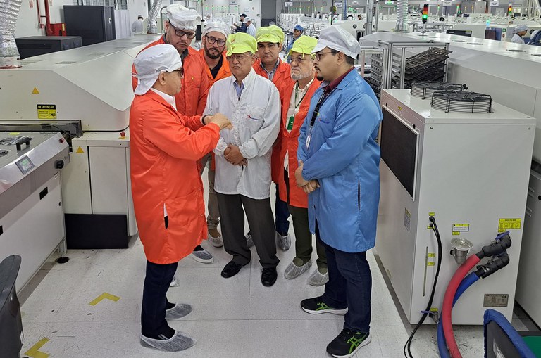 Foto mostra homem de jaleco laranja e boné branco fazendo uma explicação a outros homens também de jaleco e bonés 