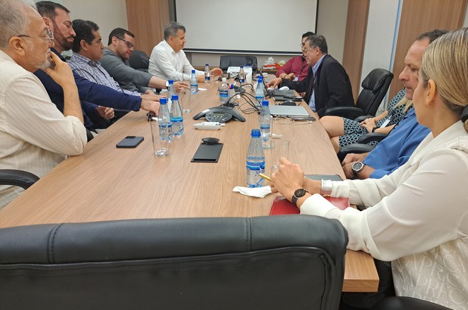 Foto mostra homem de blazer preto e de óculos em conversa com outro homem de blusa branca, observado por outras pessoas