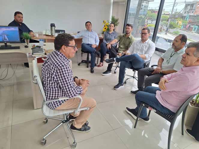 Foto mostra grupo de pessoas em reunião numa sala com um computador e uma visão externa da rua de fundo