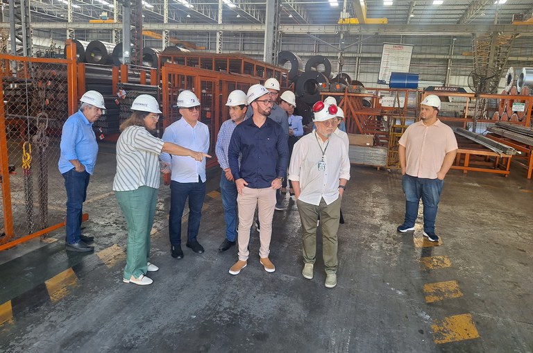 Foto mostra uma senhora de blusa listrada, calça verde e com capacete de proteção na cabeça, apontando para uma estrutura, sob os olhares atentos de um grupo de pessoas também com capacete