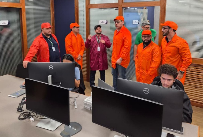 Foto mostra homens de jaleco e boné visitando uma sala com pessoas e computadores
