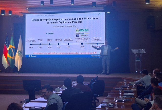 Foto mostra homem de paletó cinza fazendo uma apresentação, com um grande painel eletrônico ao fundo