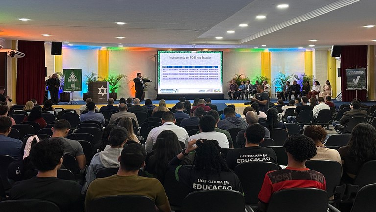 Foto mostra homem falando em evento público com um telão ao fundo