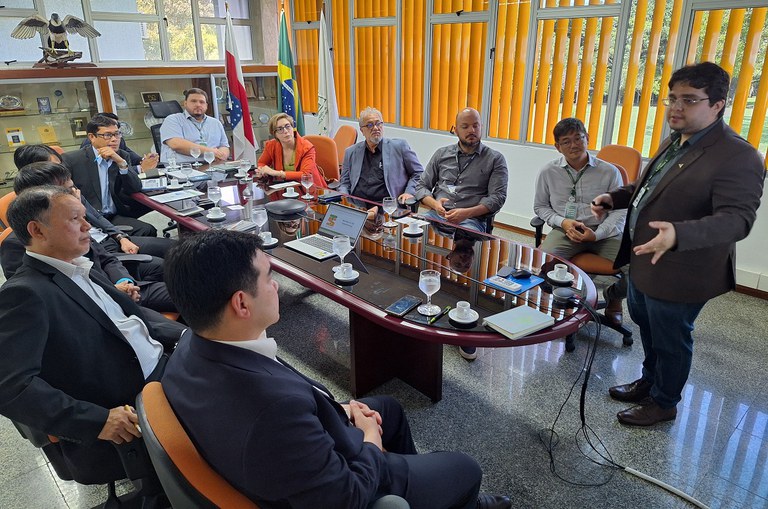 Foto mostra homem de paletó marrom em pé, explanando durante reunião com outros homens em uma sala fechada 