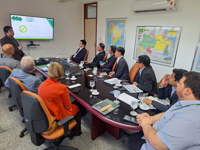Foto mostra um homem de paletó marrom em pé, explanando durante reunião com outros homens em uma sala fechada