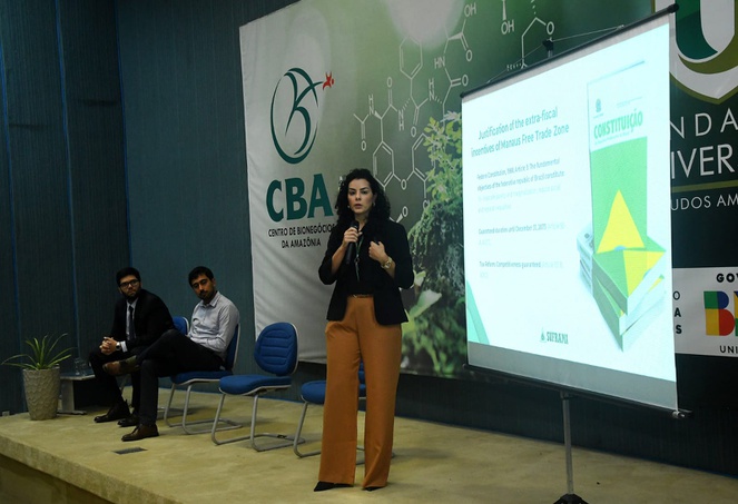 Foto mostra uma mulher em pé falando ao microfone, com dois homens observando e um telão como apoio