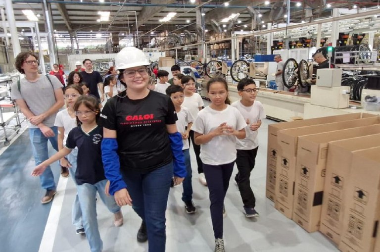 Foto mostra mulher de capacete mostrando peças de bicicletas a grupo de estudantes