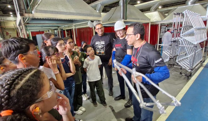 Foto mostra homem de blusa escura com mangas azuis e óculos, mostrando uma peça de bicicleta a um grupo de estudantesteção