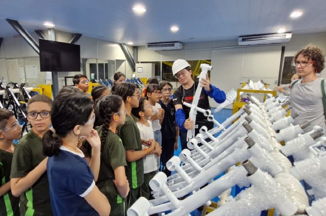 Foto mostra mulher com capacete branco e mostrando uma peça branca de bicicleta a um grupo de estudantes