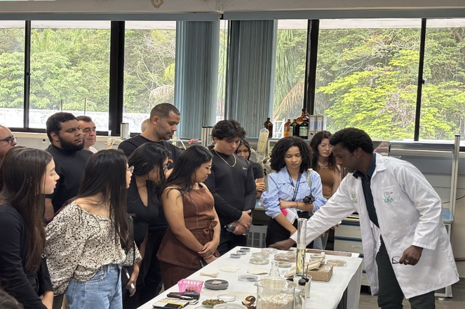 Foto mostra homem de jaleco branco mostrando produtos de pesquisa em laboratório para grupo de pessoas
