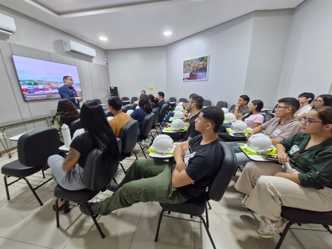 Foto pessoas reunidas numa sala ouvindo uma explicação de um homem com blusa azul e um telão como apoio