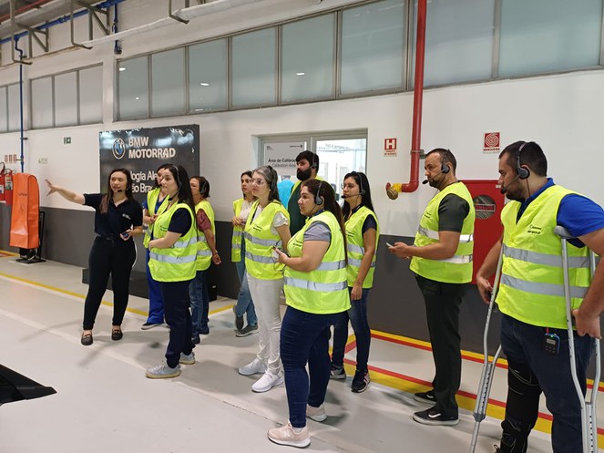 Foto mostra mulher de roupas escuras fazendo uma apresentação a grupo de pessoas com jalecos verdes
