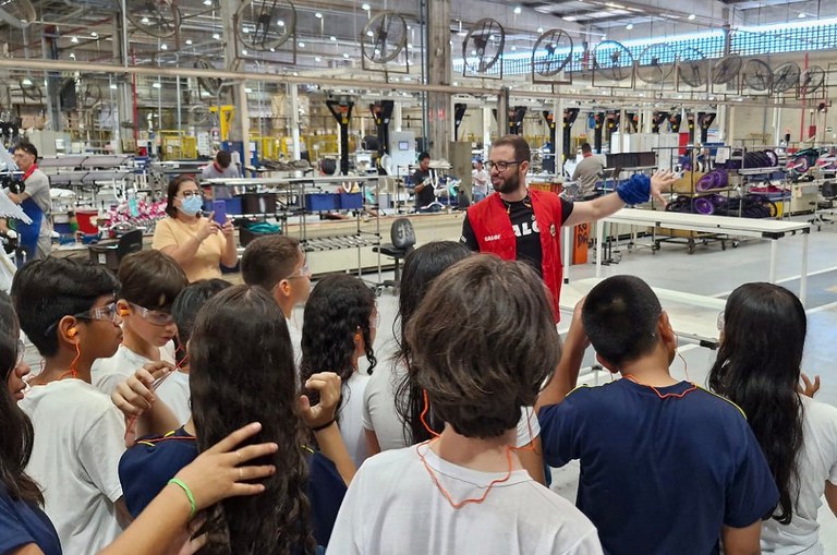 Foto mostra homem de jaqueta vermelha fazendo uma explicação a um grupo de alunos em uma fábrica de bicicletas