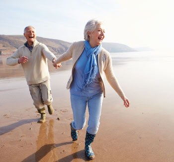 Casal de idosos correndo de mâos dadas na praia em um dia de sol.