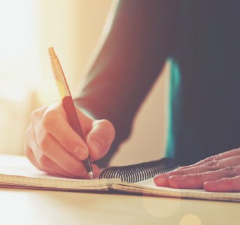 Jovem escrevendo com caneta e caderno sobre uma mesa.