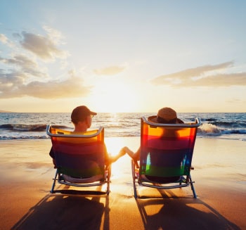 Casal sentado em cadeiras na areia da praia de frente para o mar contemplando o pôr-do-sol.