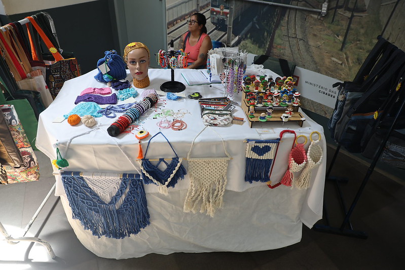 Itens de artesanato expostos em mesa e araras no saguão da Estação Mercado da Trensurb, com a artesã sentada ao fundo da imagem