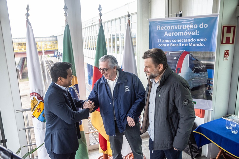 Na imagem, o secretário Marcos Daniel, o diretor-presidente da Trensurb, Nazur Garcia, e o CEO da Aerom, Marcos Coester, cumprimentando-se enquanto sorriem. Eles estão na Estação Aeroporto da Trensurb, diante de bandeiras e de um painel sobre a reconstrução do Aeromóvel.