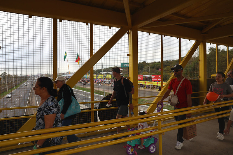 Usuários do metrô atravessam a passarela em direção à Estação Esteio do metrô; ao fundo, vê-se o parque de Exposições Assis Brasil durante a Expointer