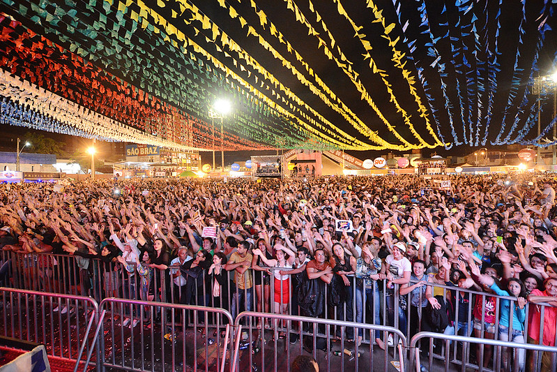 Imagem de um grande evento cultural realizado à noite, com um público numeroso reunido em espaço aberto. Em primeiro plano, milhares de pessoas estão posicionadas atrás de grades de contenção, com braços levantados, demonstrando entusiasmo e participação ativa no evento. O público é diverso, composto por pessoas de diferentes idades e perfis. Na parte superior da imagem, o céu é coberto por bandeirolas coloridas dispostas em fileiras, formando um efeito visual típico de festas populares brasileiras, especialmente festas juninas. Ao fundo, observam-se estruturas de palco, iluminação cênica, barracas e painéis com marcas e sinalizações do evento, todas iluminadas artificialmente. A cena transmite um ambiente festivo, de celebração cultural e grande participação popular, característico de eventos tradicionais e manifestações culturais de grande porte no Brasil.