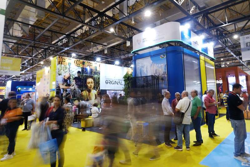 Imagem de um pavilhão de feira ou exposição realizado em ambiente interno, com estrutura metálica aparente no teto e iluminação intensa. Em destaque, observa-se um estande institucional de grande porte, com painéis verticais nas cores azul, verde e amarelo, associados à identidade visual do Governo Federal e à promoção do turismo brasileiro. Ao redor do estande, há grande circulação de visitantes, alguns em movimento, criando efeito de leve desfoque que indica fluxo intenso de pessoas. Grupos de participantes conversam e observam os expositores, caracterizando um ambiente de feira profissional. O piso colorido e a disposição dos estandes reforçam a organização do espaço e a dinâmica do evento, voltado à divulgação institucional, promoção de destinos e interação com o público.