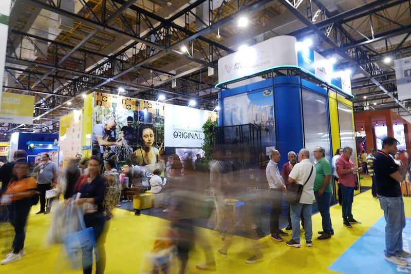 Imagem de um pavilhão de feira ou exposição realizado em ambiente interno, com estrutura metálica aparente no teto e iluminação intensa. Em destaque, observa-se um estande institucional de grande porte, com painéis verticais nas cores azul, verde e amarelo, associados à identidade visual do Governo Federal e à promoção do turismo brasileiro. Ao redor do estande, há grande circulação de visitantes, alguns em movimento, criando efeito de leve desfoque que indica fluxo intenso de pessoas. Grupos de participantes conversam e observam os expositores, caracterizando um ambiente de feira profissional. O piso colorido e a disposição dos estandes reforçam a organização do espaço e a dinâmica do evento, voltado à divulgação institucional, promoção de destinos e interação com o público.