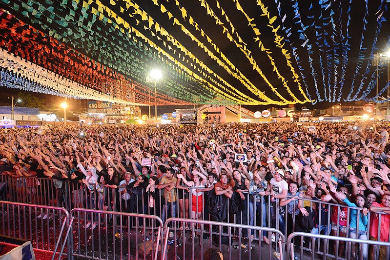 Imagem de um grande evento cultural realizado à noite, com um público numeroso reunido em espaço aberto. Em primeiro plano, milhares de pessoas estão posicionadas atrás de grades de contenção, com braços levantados, demonstrando entusiasmo e participação ativa no evento. O público é diverso, composto por pessoas de diferentes idades e perfis. Na parte superior da imagem, o céu é coberto por bandeirolas coloridas dispostas em fileiras, formando um efeito visual típico de festas populares brasileiras, especialmente festas juninas. Ao fundo, observam-se estruturas de palco, iluminação cênica, barracas e painéis com marcas e sinalizações do evento, todas iluminadas artificialmente. A cena transmite um ambiente festivo, de celebração cultural e grande participação popular, característico de eventos tradicionais e manifestações culturais de grande porte no Brasil.