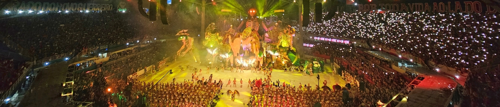 Vista panorâmica aérea de um grande evento noturno, provavelmente um desfile de carnaval ou festival folclórico como o de Parintins, em uma arena lotada (bumbódromo). A passarela central está intensamente iluminada com luzes amarelas e verdes. No centro da imagem, destaca-se uma enorme alegoria ou estrutura cênica com elementos que remetem à natureza e folhagens, incluindo uma figura imponente que parece ser o "Bicho Folharal" ou uma entidade similar. Múltiplas figuras humanas fantasiadas estão dispostas ao redor da alegoria. As arquibancadas, repletas de público, estão escuras, mas iluminadas por milhares de pontos de luz provenientes dos celulares das pessoas, criando um efeito de céu estrelado nas laterais superior direita e esquerda.