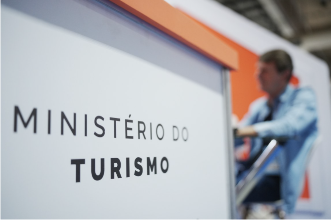 Imagem em ambiente interno que destaca, em primeiro plano, um painel institucional com a inscrição “Ministério do Turismo”, em letras pretas sobre fundo claro. O enquadramento valoriza o texto, que aparece nítido e centralizado, reforçando a identidade institucional. Ao fundo, de forma desfocada, observa-se uma pessoa sentada em uma cadeira, em contexto de evento ou espaço expositivo, sugerindo um ambiente de feira, reunião ou atividade institucional. As cores e a composição da imagem reforçam o caráter oficial e institucional da cena, associada à atuação do Ministério do Turismo em eventos públicos.