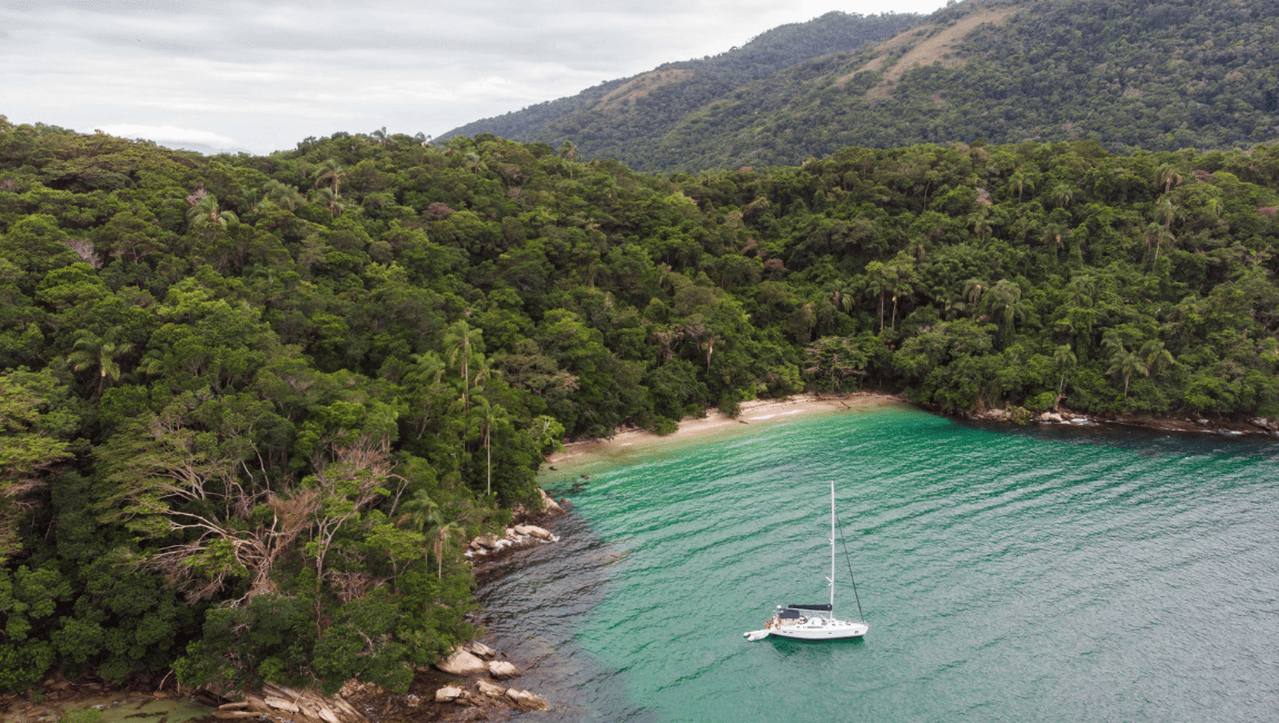 Fotografia aérea de uma baía com águas calmas e cristalinas de tom verde-azulado, onde um veleiro branco com um pequeno bote atrelado se encontra ancorado. A costa apresenta uma pequena e isolada praia de areia clara, ladeada por rochas e completamente cercada por colinas cobertas por uma densa e exuberante floresta verdejante que se estende até ao fundo sob um céu nublado.