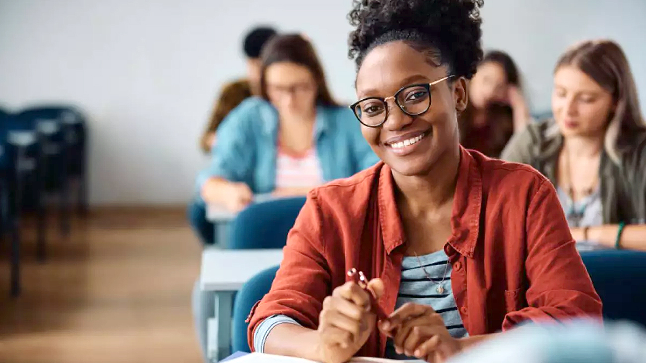 Estudante com óculos sorri para a câmera enquanto segura uma caneta em sala de aula, com outros alunos desfocados ao fundo.