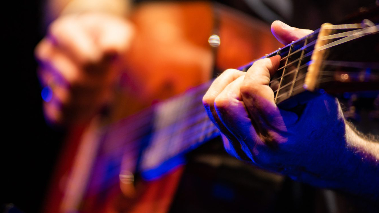 Close-up de uma mão tocando as cordas de um violão, com fundo desfocado e iluminação colorida em tons quentes e azulados.
