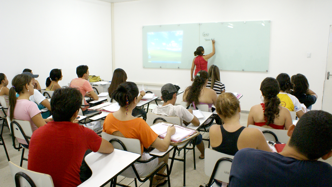 Sala de aula com estudantes sentados em carteiras enquanto uma professora escreve em um quadro branco na frente da turma.