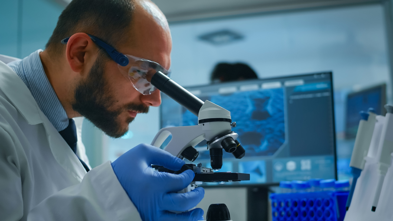 Profissional com jaleco, luvas e óculos de proteção observa amostra em microscópio dentro de laboratório.