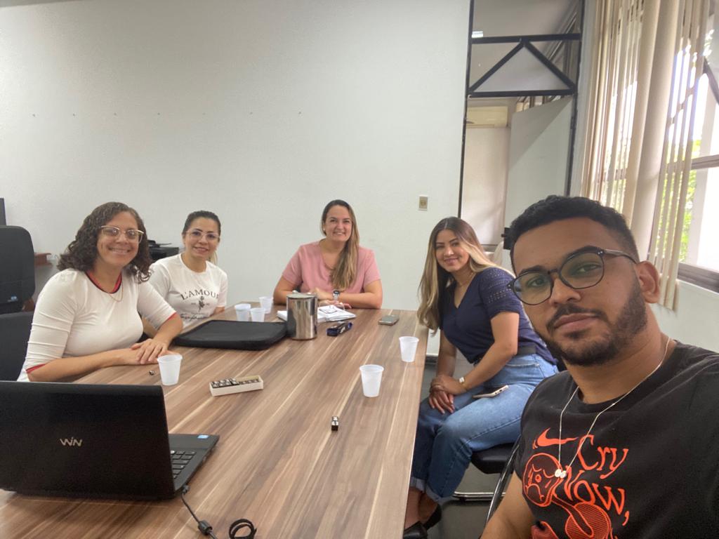 5 mulheres e um homem sentados á mesa sorrindo com notebook sobre ela..
