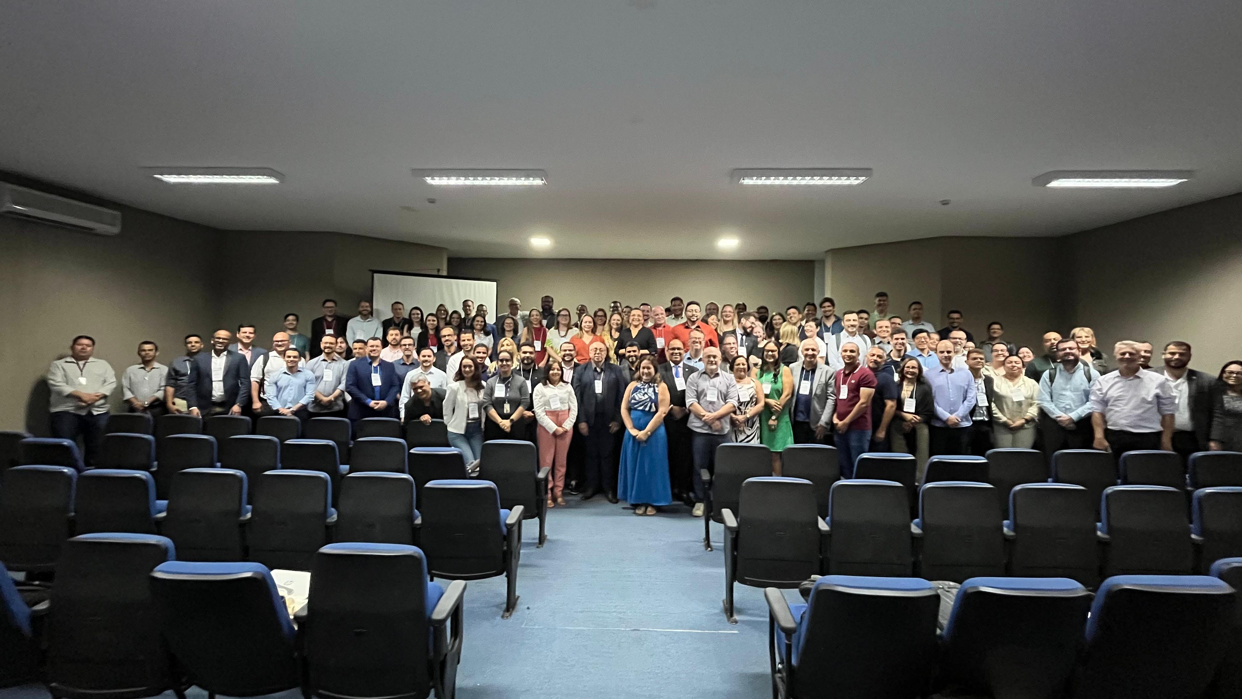 Fotografia de um grande grupo de participantes da reunião da SEPLAN reunidos à frente de um auditório, com fileiras de poltronas azuis vazias em primeiro plano.