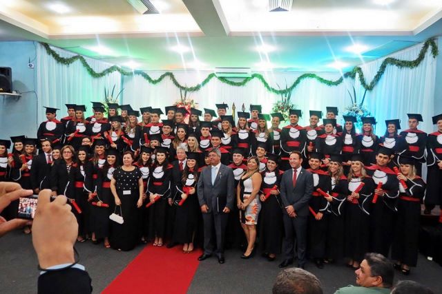 Uma turma de formandos, vestindo becas e capelos, posa para uma foto oficial em um palco decorado com guirlandas de folhas, acompanhados por autoridades ou professores no centro